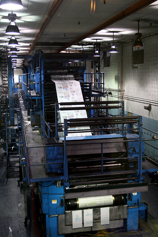 A printing press fills a long room, rolling out large sheets of printed newspaper.