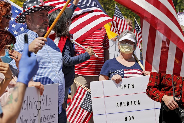A photograph of a street view showing several protestors wearing red, white, and blue clothes; one woman near the center of the image wearing a medical mask, sun visor, and holding a sign that reads, "Open American Now." Others are seen waving large American flags and holding other protest signs.