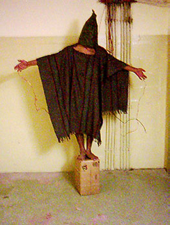 صورة فوتوغرافية تظهر شخصًا يقف على صندوق بذراعيه مرفوعتين. يتم تغطية الشخص بملابس تشبه الشال وغطاء كامل يغطي الوجه بالكامل.