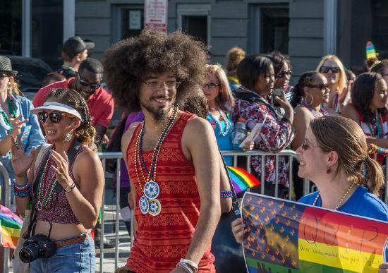 Individuals wearing bright clothes and beads smiling as they carry rainbow flags down the street