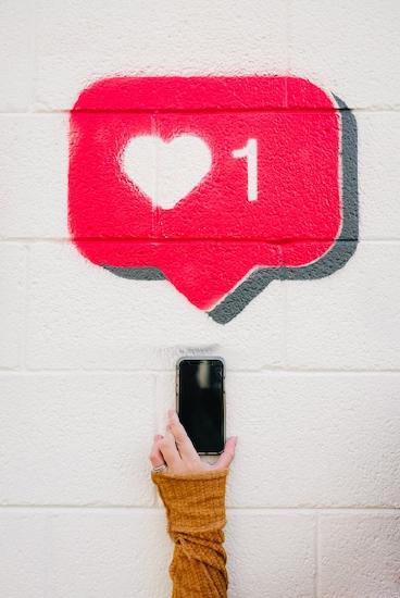 Person holding a mobile phone against a white wall, with a red heart above the phone, similar to a "like" emoji on Facebook or Instagram. Photo by Kelly Sikkema on Unsplash.