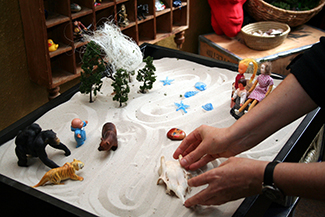 تُظهر هذه الصورة شخصًا يلعب بأشياء في صندوق صغير مملوء بالرمل. يقوم الشخص بتنظيم هذه الأشياء وشخصيات اللعب الصغيرة في شكل من أشكال العلاج يسمى اللعب بالرمل.