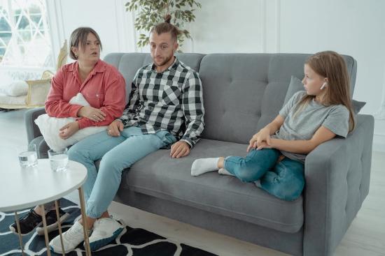 Child and parents sitting on a gray couch. Child has wireless ear buds in while parents are trying to talk to her.