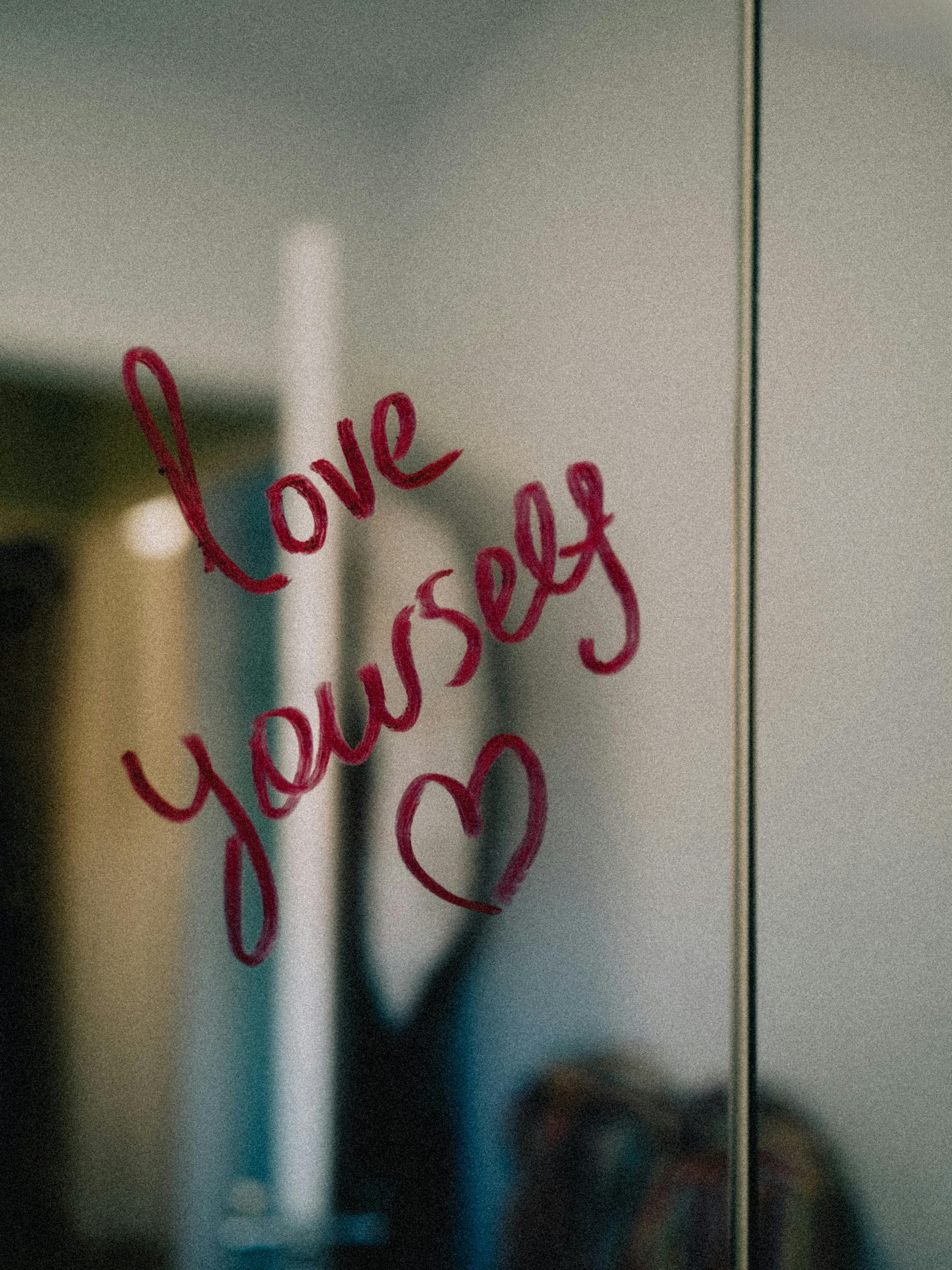 Red lipstick message "Love yourself" with a heart drawn on a mirror. The background is softly blurred, creating a warm and encouraging tone.