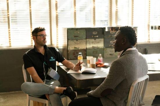 Two men sit at a table in a sunlit room having a serious conversation. A notepad and beverages are on the table, creating a focused, calm atmosphere.