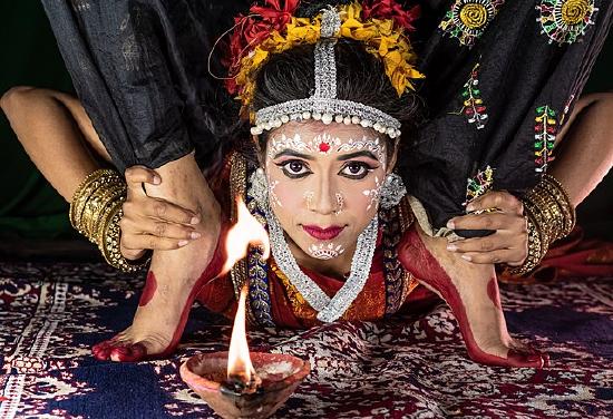 A woman with dramatic eye makeup and head jewelry, in a deep bend with her head between her feet, looking at the camera. 