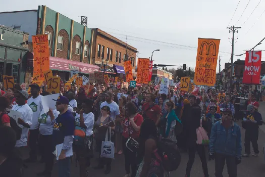 An image of a group of people marching down a street, one of whom holds a sign that reads &ldquo;I&rsquo;m not loving&rsquo; poverty wages&rdquo;.