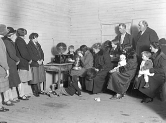 A group of women and children gather around a table with a phonograph in a bare room, listening intently.