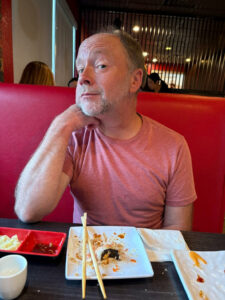A person in a pink shirt seated at a table with an empty plate. A pair of chopsticks is laid upon the edge of the plate.