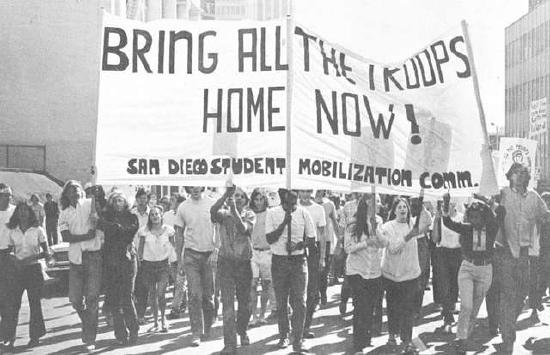 Protestors at an antiwar Vietnam march holding a sign stating bring all the troops home now