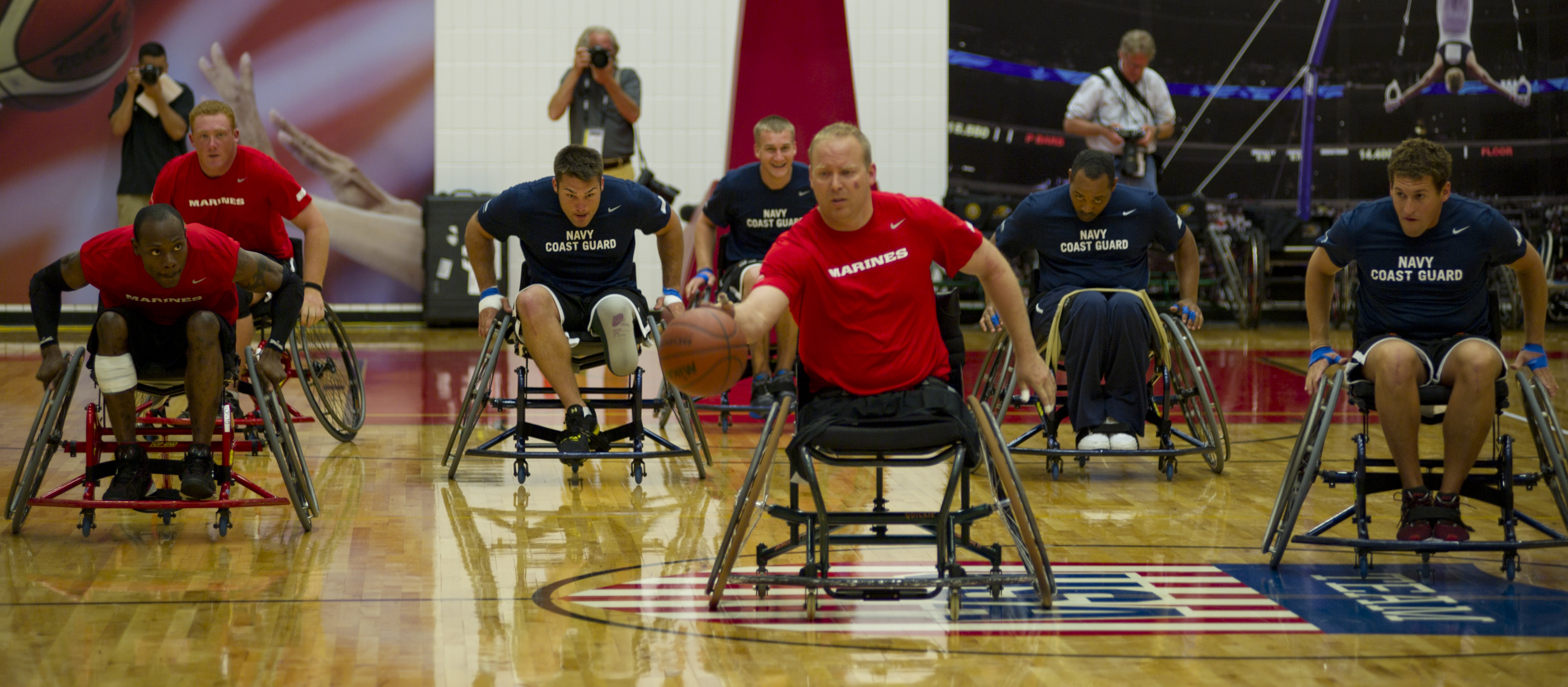 Athletes in wheelchairs compete in basketball, with several wearing team jerseys in a gym setting.