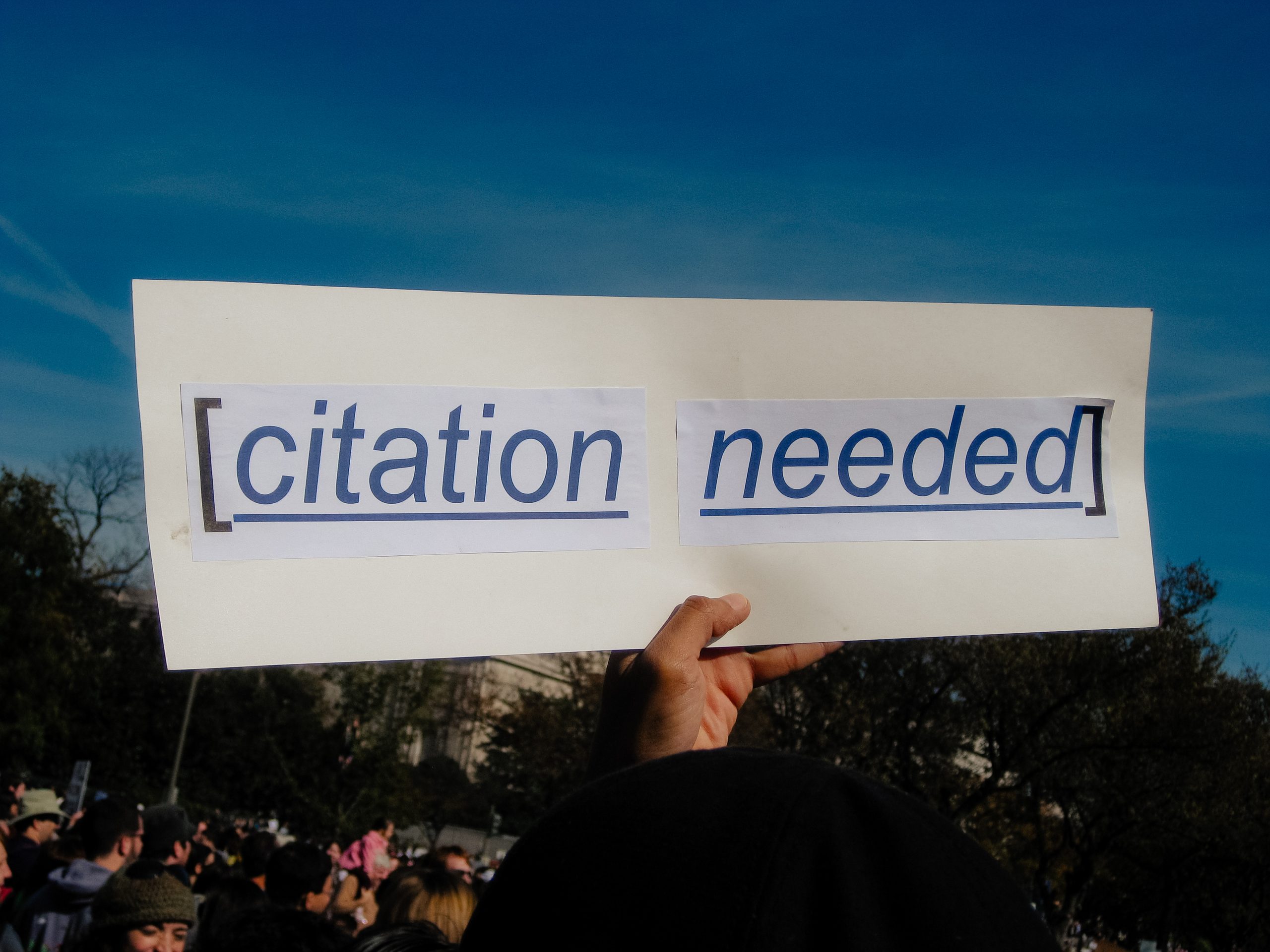 A protestor holds up a white sign with the phrase &ldquo;[citation needed]&rdquo; printed in bold blue text, mimicking the style of Wikipedia&rsquo;s citation prompt. The background shows a crowd of people gathered outdoors with trees and a clear blue sky overhead.