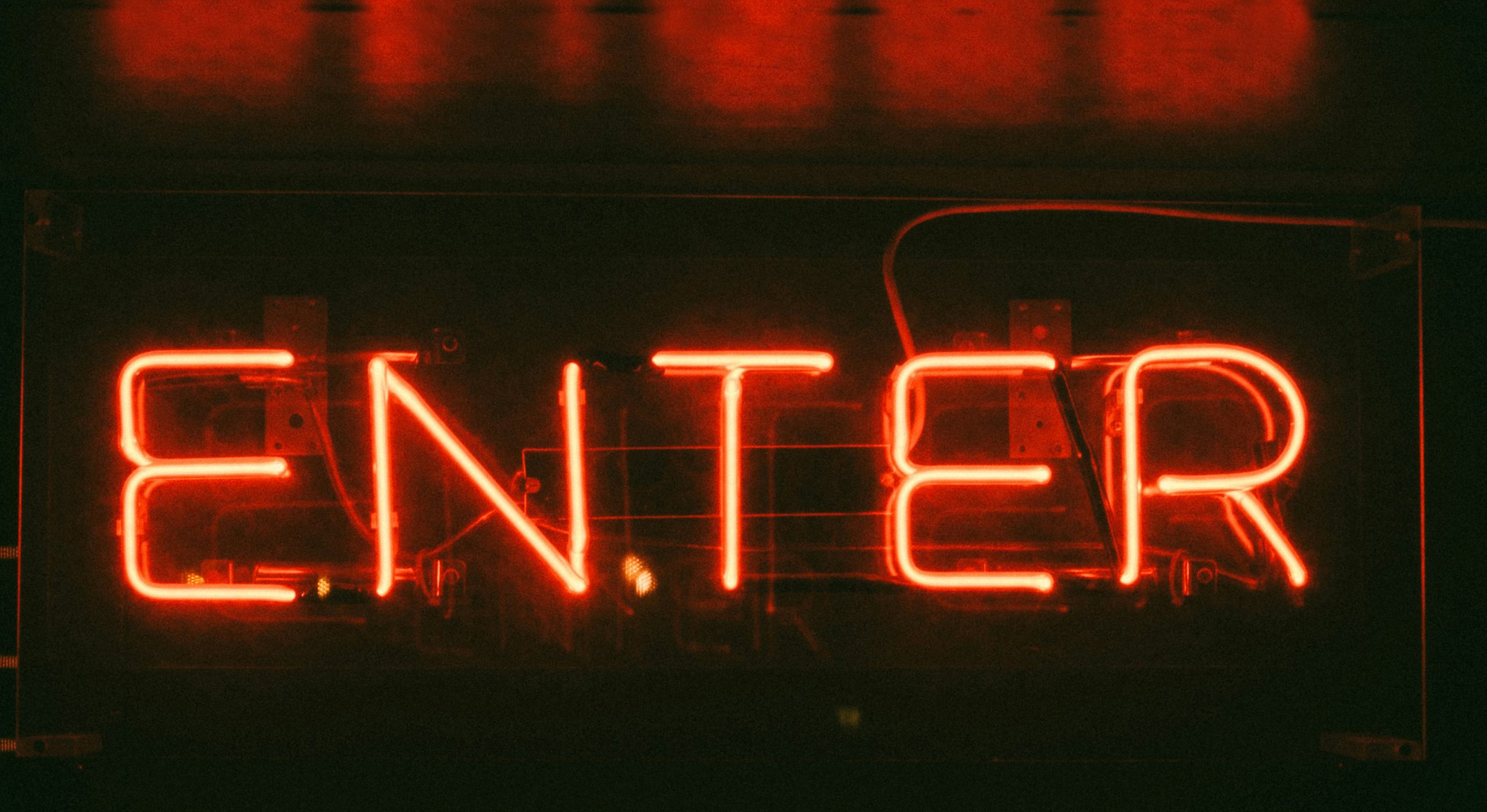 A glowing red neon sign displaying the word "ENTER" in capital letters, set against a dark background. The light from the sign casts a warm red hue around it, creating a moody, inviting atmosphere often seen at bars or clubs.