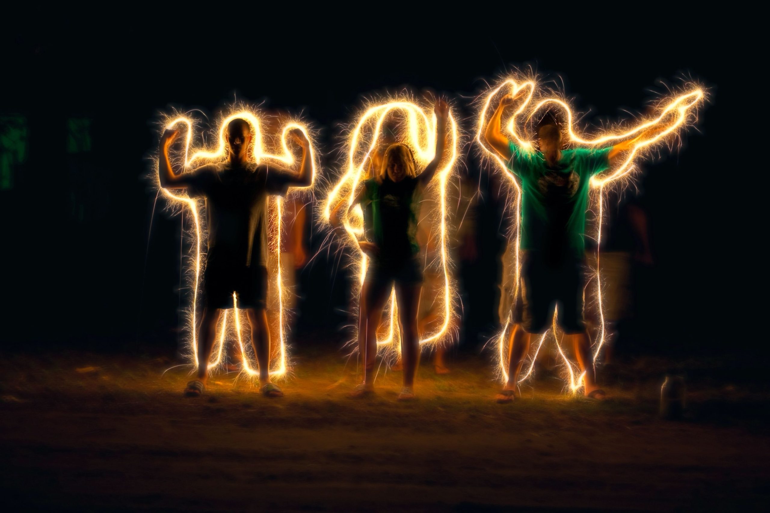 Four people pose in the dark while sparkler trails outline their bodies, creating glowing, electric-like silhouettes. The long exposure photo captures the motion of the sparklers, producing a vibrant and playful energy effect against the night backdrop.