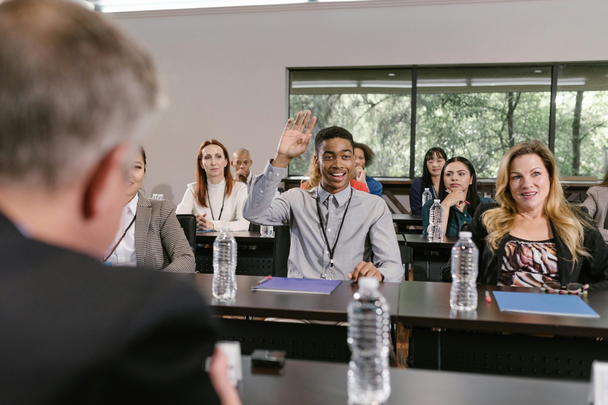 A young man sitting in a conference or classroom setting enthusiastically raises his hand, smiling as he engages with a speaker or presenter. Around him, other attendees of various ages and backgrounds listen attentively, seated at tables with water bottles, notepads, and lanyards. The atmosphere appears professional and interactive, with large windows in the background providing a view of greenery.