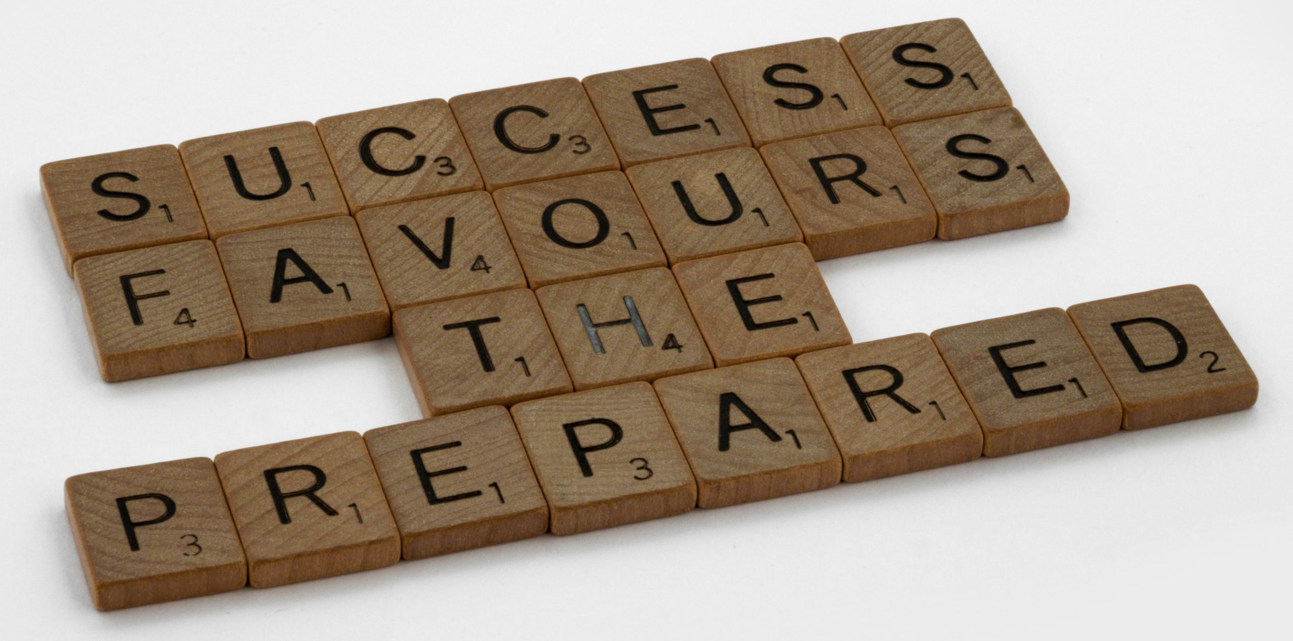 Scrabble-style wooden letter tiles spell out the phrase &ldquo;SUCCESS FAVOURS THE PREPARED&rdquo; on a white background. Each tile includes a point value, emphasizing a motivational message through a classic word game format.