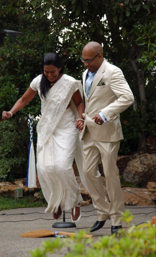 A couple with a dark skin tone jumping over a broom at their wedding.