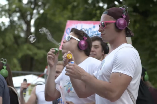 two men in white t-shirts, sunglasses, and purple headphones blow bubbles while standing in a crowd carrying signs.