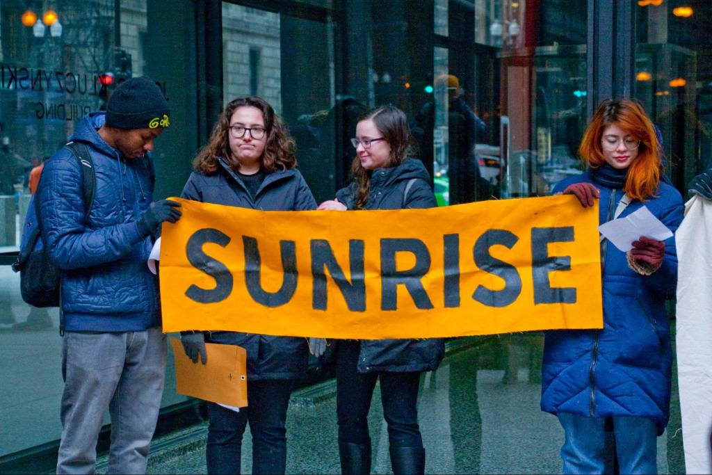 three young adults stand in the street with warm clothing, holding an orange and blue banner that reads, "Sunrise."