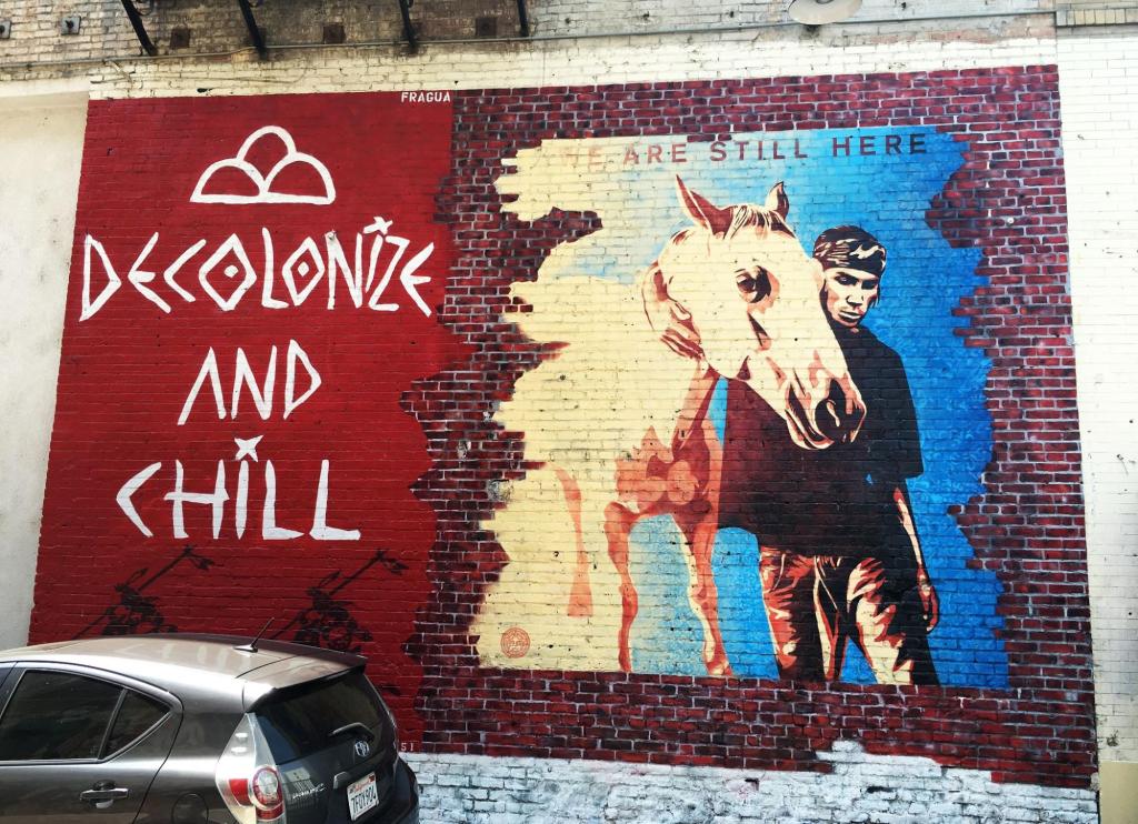 a mural painted on brick with a horse, and casually dressed man beside it. Text reads, decolonize and chill."