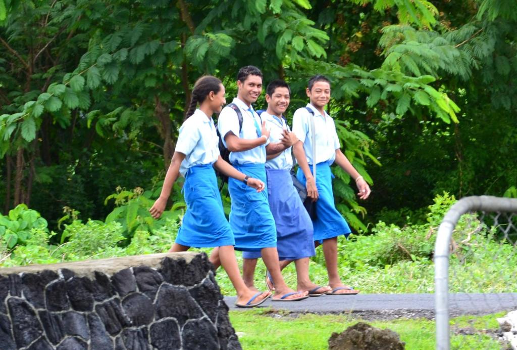 four teenagers in straight blue skirts walking on a trail with lush forest behind them.