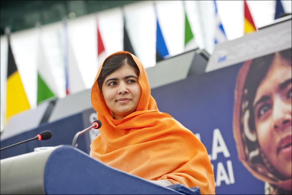 Malala Yousafza&iuml; wearing a bright orange head scarf standing at a podium in front of her image on a billboard.