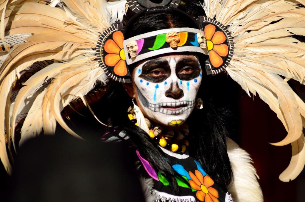 a profile of a person with an elaborate headdress with feathers, a colorful headband with flowers, and a painted face in black and white.