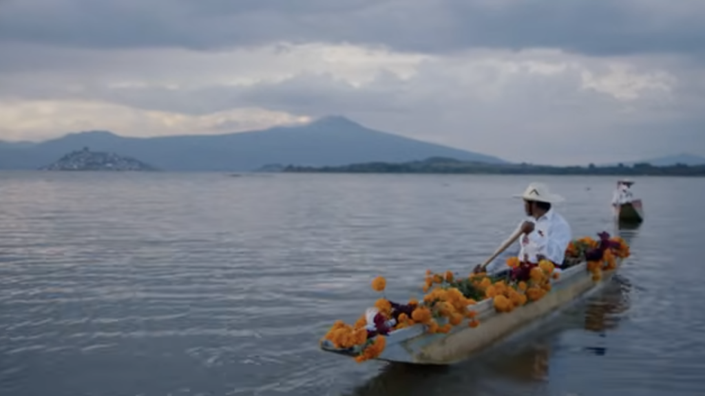 two narrow boats are rowed by people, moving gently in a lake, with mountains below. the boat in the foreground carries many orange flowers