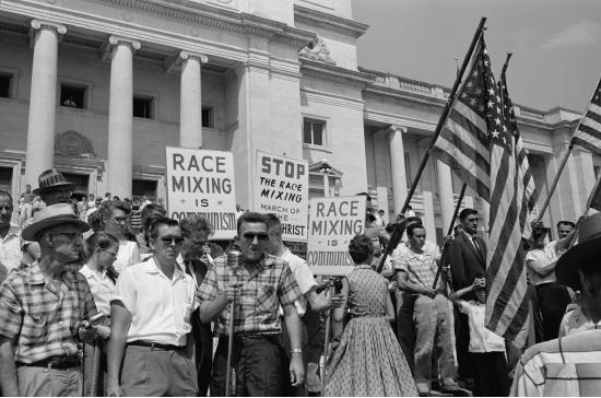 	Little Rock, 1959. Rally at state capitol, protesting the integration of Central High School. Protesters carry US flags and signs reading "Race Mixing is Communism" and "Stop the Race Mixing March of the Anti-Christ".