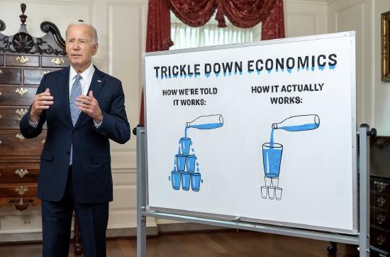 Joe Biden standing beside a whiteboard labeled &ldquo;Trickle Down Economics,&rdquo; showing two diagrams of water pouring from a bottle into stacked cups to illustrate how economic benefits are said to flow through society.