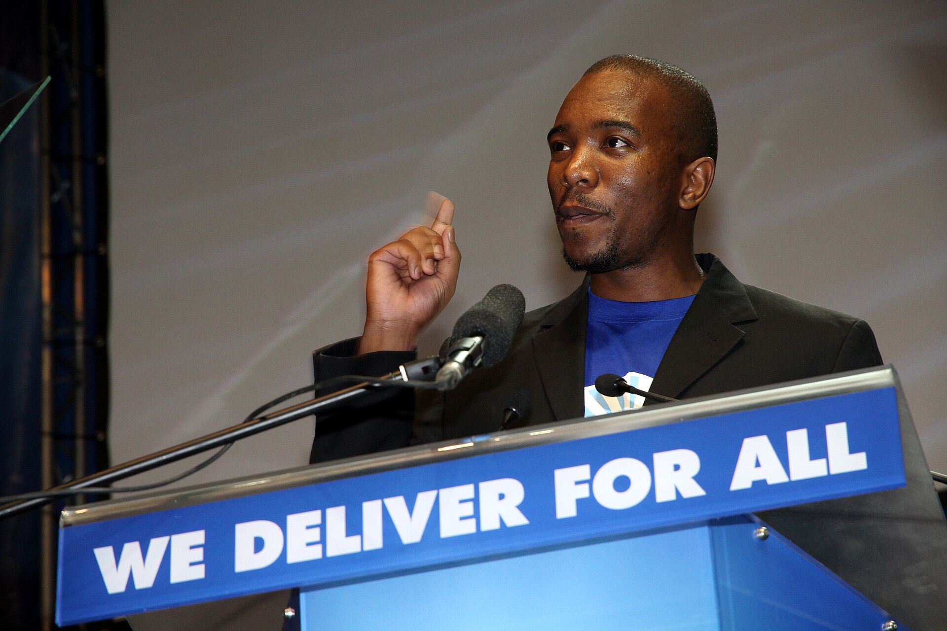 Mmusi Maimane, a South African politician, stands at a podium speaking into a microphone with his right hand lifted in a gentle fist. The phrase, "We Deliver For All" is displayed on the front of the podium. 