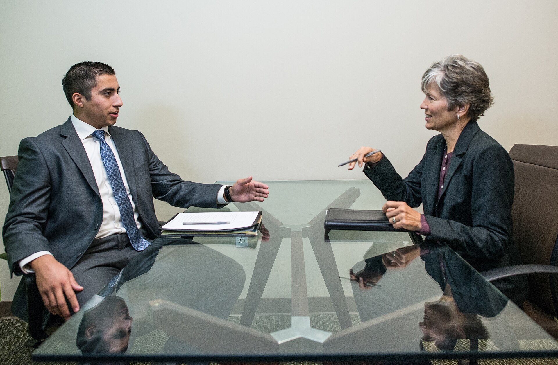 A young man seated in an office during a job interview, facing an older woman interviewer across a desk in a professional setting.