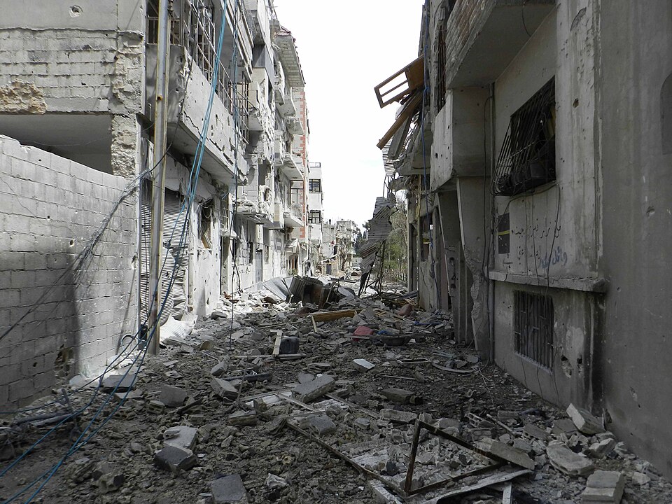 Photograph from Homs, Syria of a street littered with rubble and surrounding buildings partially destroyed from bombings.
