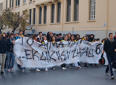 Voici une photo de jeunes qui défilent dans une rue avec une pancarte avec des inscriptions grecques dessus.