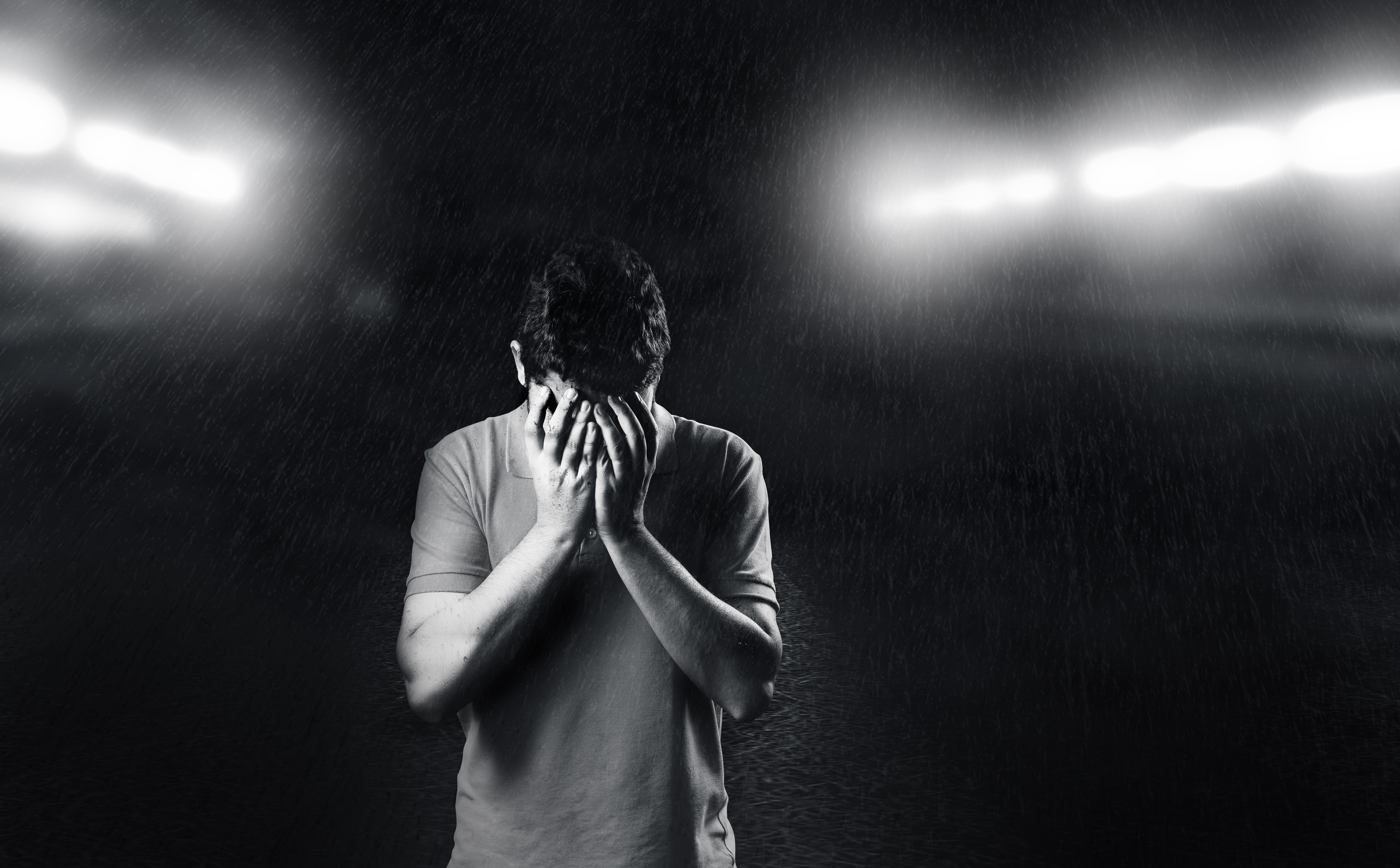 A man stands with his face covered by his hands against a black background with bright lights.