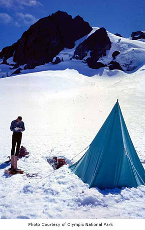 Un homme est représenté en train de prendre des notes à l'extérieur d'une tente dans les montagnes.
