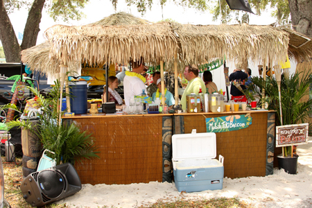 Plusieurs personnes portant des t-shirts et des leis colorés sont montrées en train de parler et de boire dans un bar tiki en plein air.