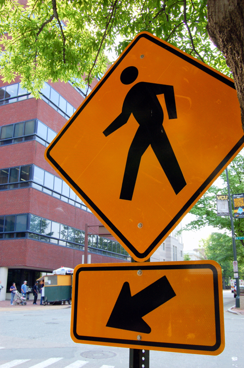 A foto (a) mostra um sinal de uma passagem para pedestres e uma flecha.