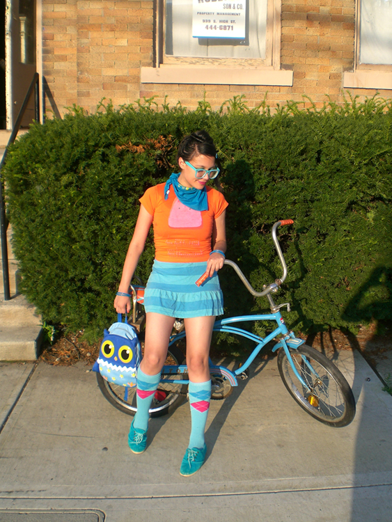 Une jeune femme vêtue de vêtements aux couleurs vives et portant un sac à main en forme de hibou est représentée debout devant un vélo bleu vintage, une grande haie et une maison de ville.