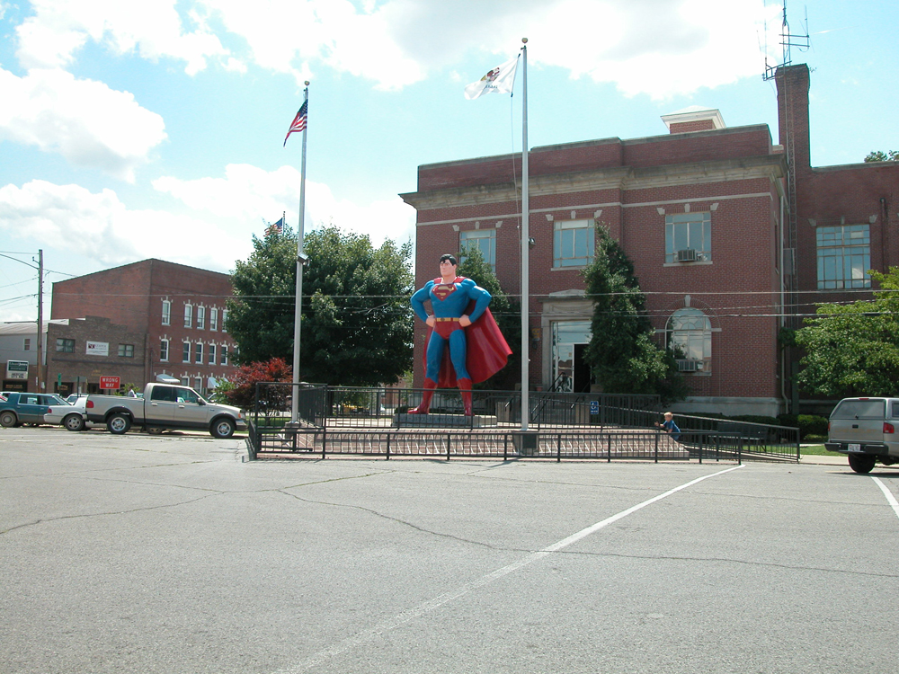 Se muestra una estatua de Superman entre dos astas de bandera y frente a un edificio de ladrillo de dos pisos.