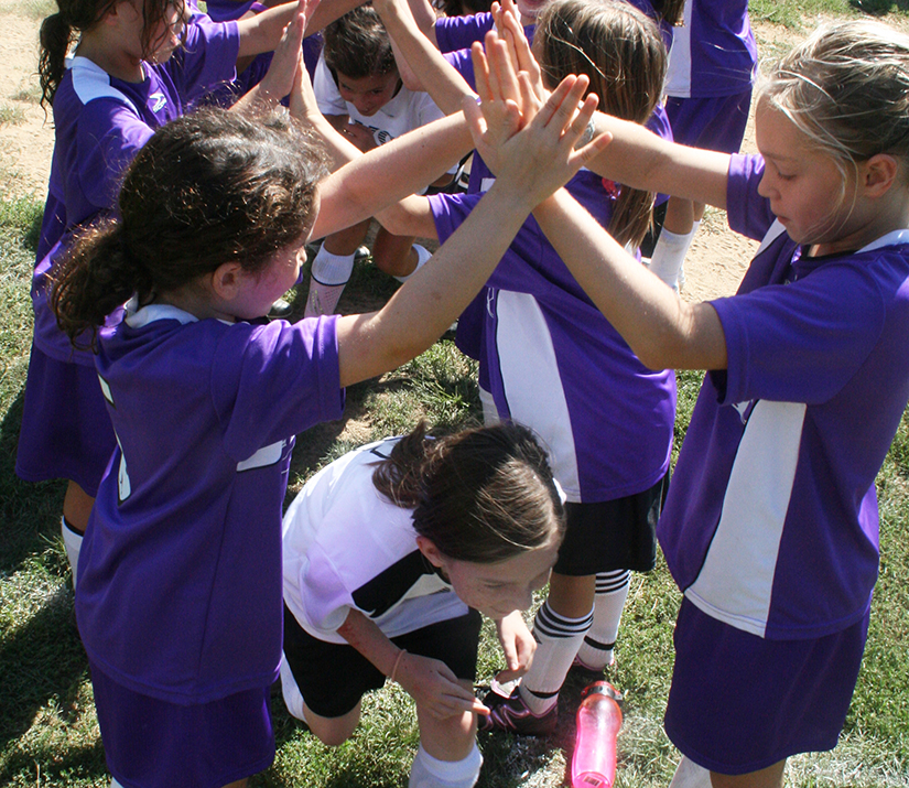 Una foto de chicas jóvenes vestidas con uniformes de fútbol formando un túnel con las manos por el que otras chicas corren como ritual posterior al juego.
