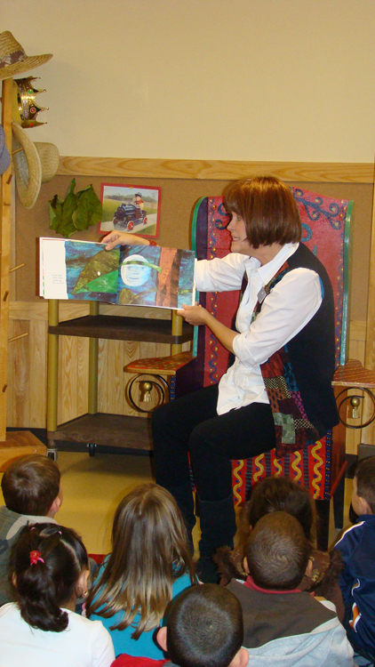 Une enseignante est représentée assise sur une chaise et lisant un livre d'images à un groupe d'enfants assis par terre devant elle.