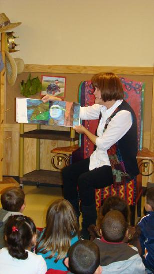 A female teacher is shown sitting in a chair and reading a picture book to a group of children sitting in front of her on the floor.