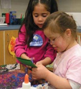 Two girls wearing pink and making crafts at a table. One of the girls is helping the other to cut a green piece of paper.