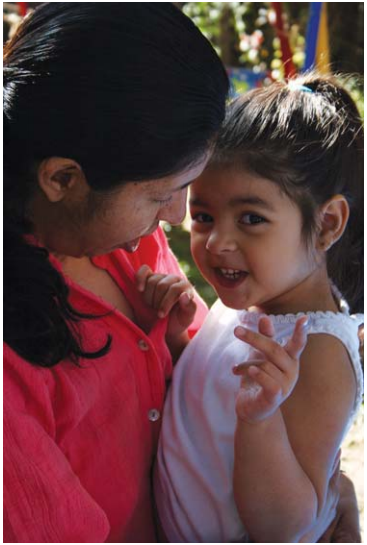 A woman holding a toddler girl and looking down at her while the toddler smiles and points upward, looking at the camera.