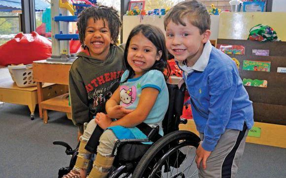 Dos niños sonriendo a la cámara y de pie a ambos lados de una niña en silla de ruedas. Ella también está sonriendo y mirando a la cámara.