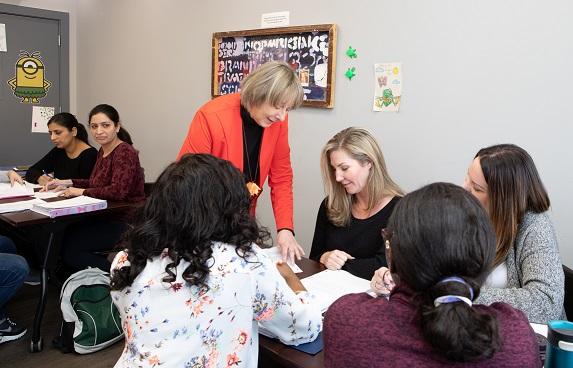Un aula de mujeres aprendiendo a ser maestras. El instructor se encuentra en una mesa con un grupo de alumnos, platicando con ellos.
