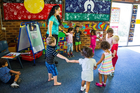 Un maestro cogido de la mano con un círculo de jóvenes estudiantes. Están parados en un aula con decoraciones hechas para niños.