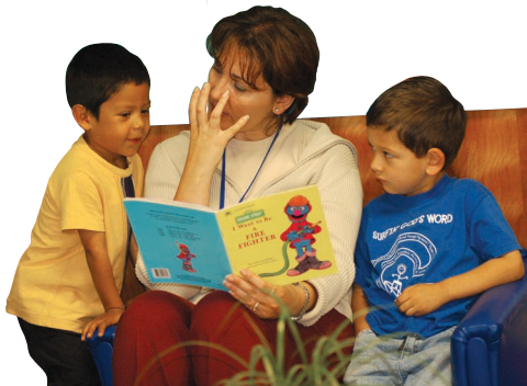 Una maestra leyendo a dos chicos sentados a su lado. La maestra está interpretando algo del libro al colocar su mano en forma de garra cerca de su boca.