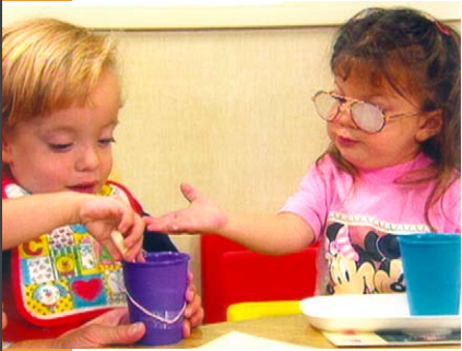 Two children with disabilities sitting at a table. The boy is placing something inside a purple cup and the girl is gesturing for it with her hand.